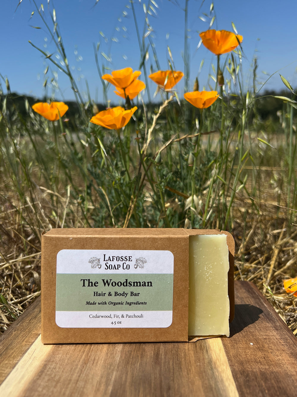 Bar of soap with 'The Woodsman' label in a natural setting with orange flowers and grass.