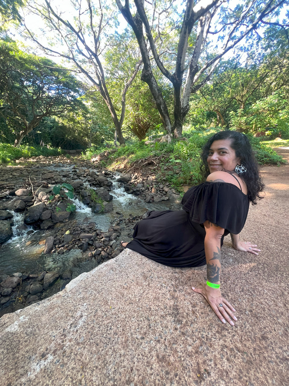 Sabrina in a black dress sitting on a rock by a stream with trees in the background